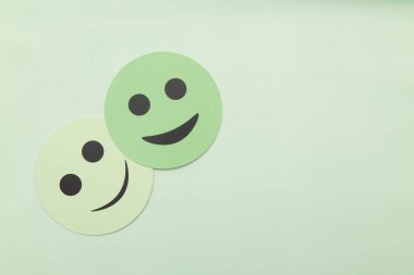 Açık yeşil arka planda fotokopi alanı olan iki yeşil kağıt kesiği gülen surat. Zihinsel sağlık anlayışı, olumlu geri bildirim, müşteri memnuniyeti ve minimalist tarzda duygusal mutluluk kavramı.