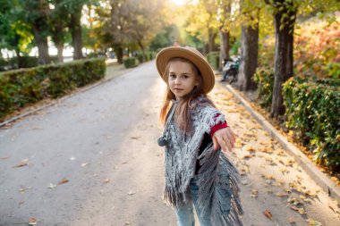 Hareket özgürlüğü. Küçük kız 7 yaşında, sıcak bir pelerinli ve şapkalı. Sonbaharda günbatımında parkın arka sokağında dans ediyor.