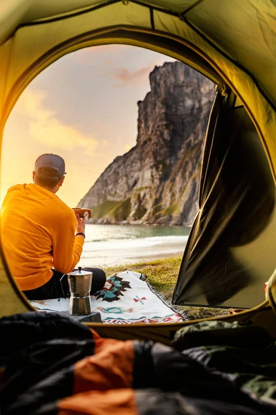 Günbatımında, Norveç 'in kuzeyinde, Lofoten adalarında, Kvalvika sahilinde çadırda. Çadırın önünde elinde sıcak içecek bardağıyla oturan bir adam..