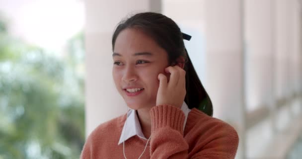 長髪の可愛いアジア系高校生のスローモーションシーン 携帯電話で白いイヤホンを使った音楽を聴く — ストック動画