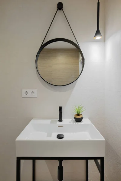 Interior de baño de estilo moderno en apartamento reformado. Fregadero blanco con grifo negro y marco de espejo redondo, iluminado por una lámpara. —  Fotos de Stock