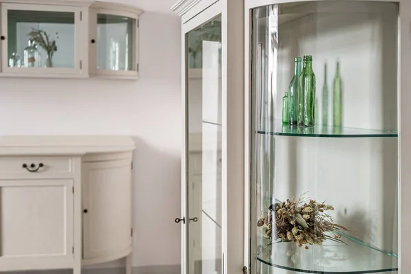 Weiße Schränke im alten Stil mit Glasregalen in einer Wohnung im klassischen Stil. Grüne Glasflaschen und getrocknete Blumen zur Dekoration. — Stockfoto