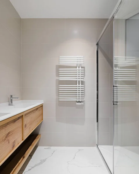 Modern fürdőszoba fa alappal a fehér mosogatókhoz. Hűtőszekrény meleg vizes eljárásokhoz télen. Zuhanyzóna üvegajtóval. Könnyű csempével díszített fürdőszoba. Stock Kép