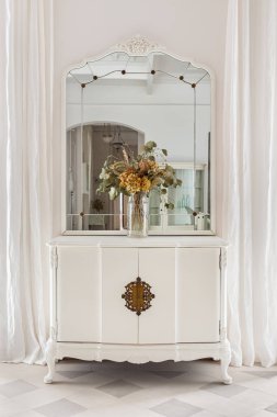 Ahşap dolapta klasik ayna ve çiçek buketi. Beyaz duvar ve perdelerle klasik parlak iç dekorasyonlu eski mobilyalar.. .