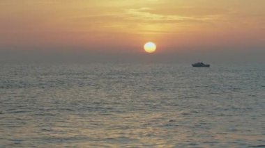 Güneş doğarken küçük, yalnız bir balıkçı teknesinin silueti yelken açıyor. Mavi denizin üzerinde yükselen güneşli altın gökyüzü. Okyanus kıyısında yumuşak renk günbatımı, güzel deniz manzarası.