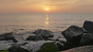 Güneş denizin üzerinde yükseliyor. Güneş doğarken sahilde kayalara vuran dalgalar ve arka planda bir tekne.