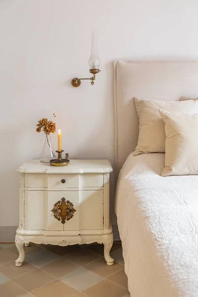 Klasyczna sypialnia z drewnianym stołem nocnym z płonącą świecą i kwiatami w pobliżu wygodnego łóżka. Wnętrze przytulnego domu w stylu retro. — Zdjęcie stockowe