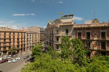Barselona 'da canlı bir sokak sahnesi. Tarihi binalar, yeşil ağaçlar ve açık mavi gökyüzü. Arabalar yol boyunca park edilmiş, şehir yaşamını gözler önüne seriyorlar..