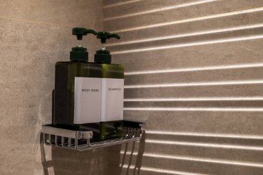 Metal bir duş rafında etiketli iki yeşil pompa şişesi Vücut Yıkama ve Şampuan. Yumuşak ışık duvarda çizgili gölgeler yaratır. Modern banyo ayarları.