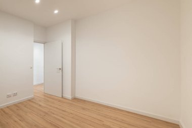 Beyaz duvarları ve ahşap zemini olan boş bir oda. Sol tarafta iki kapı görünüyor. Parlak ışıklandırma geniş hissi arttırır.