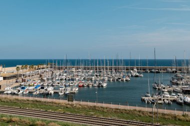 Barcelona 'da çok sayıda yelkenlisi olan bir yat limanı. Açık mavi gökyüzü sakin suyu yansıtır. Yeşil çimenler ve tren yolları yakınlarda görünüyor..