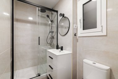 Modern banyo içinde cam duş, beyaz lavabo ve tuvalet var. Tarafsız fayanslar ve bir pencereden gelen doğal ışık temiz, minimalist bir alan yaratır..