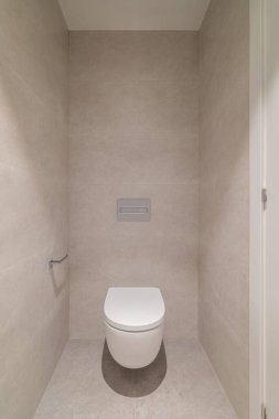 Açık gri fayanslı bir duvara karşı beyaz bir tuvaletin kullanıldığı modern, minimalist bir banyo. Alan temiz ve dağınık. Basitliği vurguluyor..