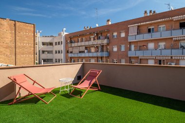 Barcelona 'da güneşli bir teras, iki kırmızı şezlong ve yapay çimlerin üzerinde küçük bir masa. Çevredeki binalar arka planda görünür..