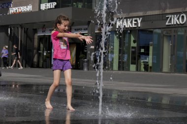 Şehrin sokaklarında, sıcakta bir fıskiyede su püskürten bir kız. Yaz tatili kavramı ve yazın anormal sıcak. Mogilev, Belarus - 21 Haziran 2021