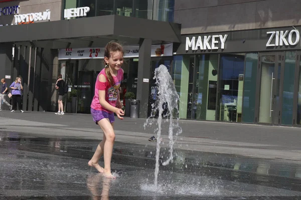 Bir kız bebek, yaz sıcağında şehir sokaklarında serinlemek için çeşmede suyla neşeyle oynuyor. Sıcak hava dalgası konsepti. Mogilev, Belarus - 21 Haziran 2021.