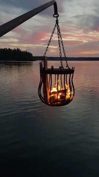 A bonfire burns in a metal basket hanging over the water at sunset