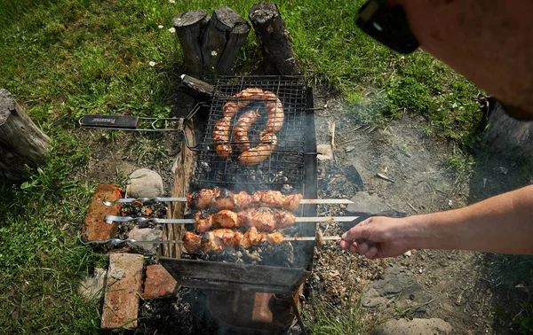 Carne de barbacoa y salchichas o salchichas en una parrilla en el patio trasero. Hombre preparando shashlik o shish kebab sobre carbón vegetal. Carne a la parrilla en pincho de metal al aire libre. Fiesta de barbacoa o picnic. Primer plano. —  Fotos de Stock