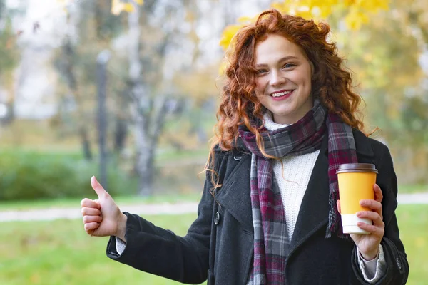 Güzel, genç, kızıl saçlı bir kadının portresi baş parmağını havada tutuyor. Ginger gülümsüyor, elinde bir bardak kahve tutuyor. Boşluğu kopyala.