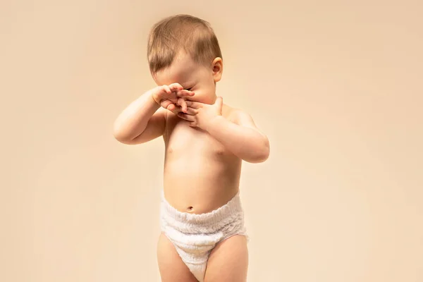 Sad little boy crying, rubbing his eyes with his hands. Allergies or tears in a baby. itchy, child itchy eyes baby wearing diapers. On a solid background monochromatic in the studio