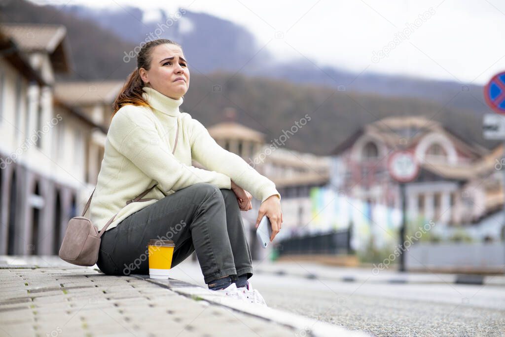 The female sits on the side of the road, on the sidewalk. Desperate woman lost in the city. Girl crying, emotion of despair