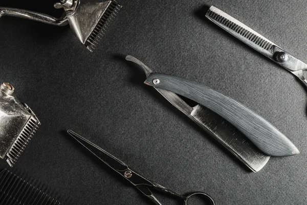 On a black surface are old barber tools. two vintage manual hair clipper, comb, razor, hairdressing scissors. black monochrome. Close-up. Barbershop background. contrast shadows. horizontal orientation. Top view, flat lay.