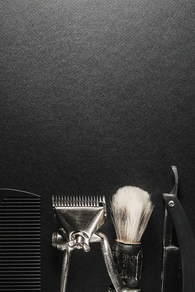 On a black surface are old barber tools.Two vintage manual hair clipper, comb, razor, shaving brush, hairdressing scissors. black monochrome. Close-up. Barbershop background. contrast shadows. Vertical orientation. Top view, flat lay. copy space
