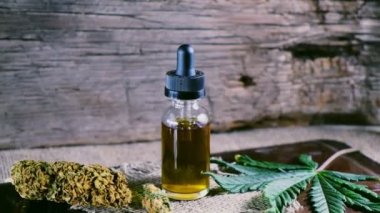 Bir şişe esrar konsantresi CBD, kurumuş marihuana tomurcukları ve tahta bir çuval kumaş arka planında kenevir yaprağının yanında duruyor. 4k