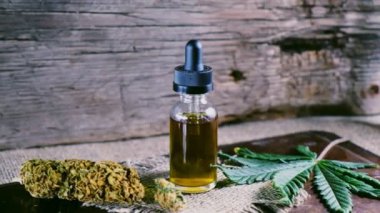Bir şişe esrar konsantresi CBD, kurumuş marihuana tomurcukları ve tahta bir çuval kumaş arka planında kenevir yaprağının yanında duruyor. 4k