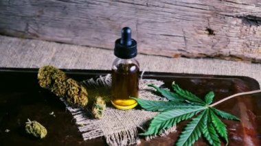 Bir şişe esrar konsantresi CBD, kurumuş marihuana tomurcukları ve tahta bir çuval kumaş arka planında kenevir yaprağının yanında duruyor. 4k