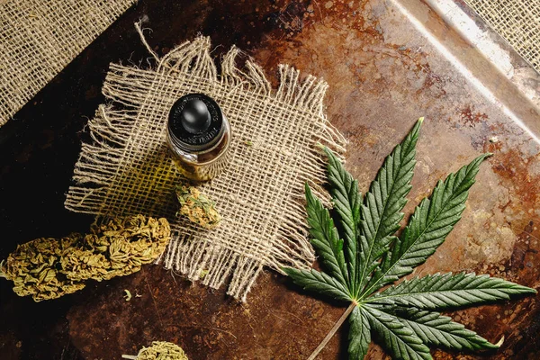 Bir şişe kenevir konsantresi, kurutulmuş marihuana tomurcukları ve paslı ahşap ve çuval bezi arka planında kenevir yaprağının yanında duruyor. Üst Manzara. Boşluğu kopyala. Yatay yönelim