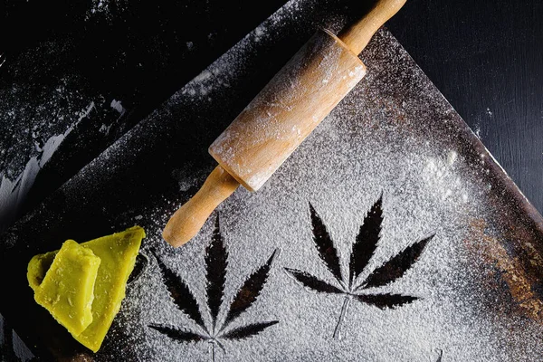 Un üzerinde marihuana yaprağı, merdane, üç kurabiye ve kenevir yağı. Paslı bir tavada. En üstteki düz yerde. Yatay yönelim