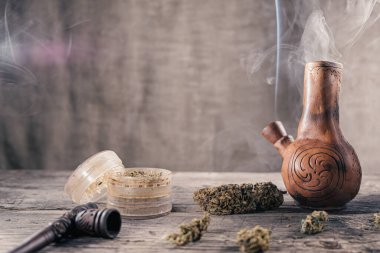 Ahşap yüzeyde tıbbi marihuana ile birlikte tüttüren eski bir kil nargilesi var. Yakınlarda tıbbi esrar tomurcukları ve öğütücü var. film tahıl etkisi. kopyalama alanı. Yatay yönelim.