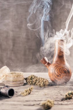 Ahşap yüzeyde tıbbi marihuana ile birlikte tüttüren eski bir kil nargilesi var. Çok duman var. Yakınlarda tıbbi esrar tomurcukları ve öğütücü var. film tahıl etkisi. Dikey yönelim