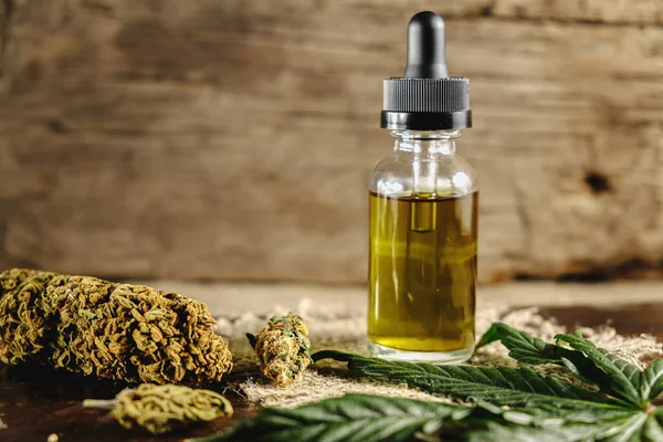 Bir şişe kenevir konsantresi, kurutulmuş marihuana tomurcukları ve paslı ahşap ve çuval bezi arka planında kenevir yaprağının yanında duruyor. kopyalama alanı. yatay yönelim