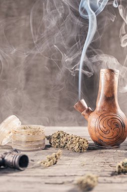 Ahşap yüzeyde tıbbi marihuanayla birlikte tüttüren eski bir balçık nargilesi var.