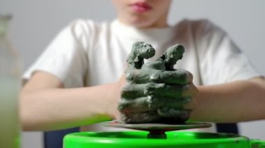Yedi yaşında, kille kirlenmiş bir çocuk çömlekçi çarkında kilden bir ürün yapmaya konsantre oluyor. Yakın plan. Yatay.