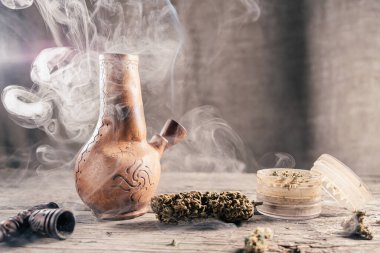 Ahşap yüzeyde tıbbi marihuana ile birlikte tüttüren eski bir kil nargilesi var. Çok duman var. Yakınlarda tıbbi esrar tomurcukları ve öğütücü var. film tahıl etkisi. kopyalama alanı. Yatay yönelim.