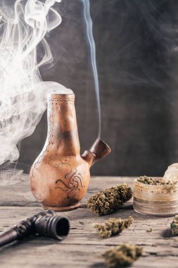 Ahşap yüzeyde tıbbi marihuana ile birlikte tüttüren eski bir kil nargilesi var. Çok duman var. Yakınlarda tıbbi esrar tomurcukları ve öğütücü var. film tahıl etkisi. Dikey yönelim