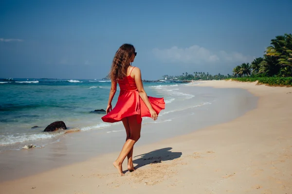 Gelukkige vrouw genieten van strand ontspannen vreugdevolle in de zomer door tropische blauwe water. Mooie rode jurk model gelukkig op reizen dragen strand zon stro hoed aan zandstrand — Stockfoto