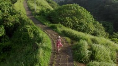 Campuhan Tepesi 'nde yürüyen kırmızı elbiseli ve hasır şapkalı genç bir kadının üzerinde uçan hava aracı. Sanatçılar, Bali' de yeşillik yolu, Ubud..