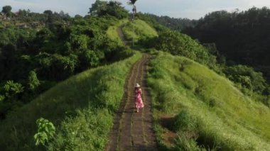 Kırmızı elbiseli ve hasır şapkalı genç gezgin kadın boş Campuhan Tepesi yolunda koşuyor. Sanatçılar, Bali 'deki yeşil yol, Ubud, Hava Aracı manzarası. 