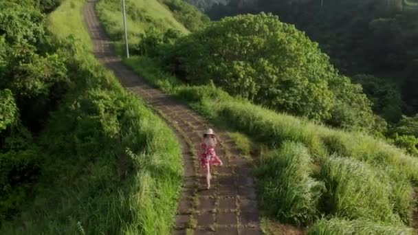 Avión no tripulado volando sobre Mujer viajera joven en vestido rojo y sombrero de paja caminando en Campuhan Ridge camino de los artistas, camino verde en Bali, Ubud. — Vídeos de Stock