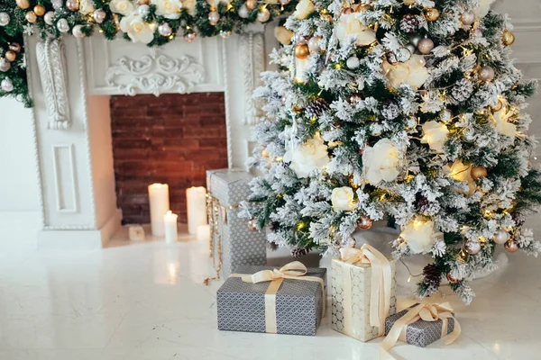 Stos prezentów świątecznych w białym i szarym papierze pod choinką ozdobione białe i złote kulki w pobliżu kominka w salonie. — Zdjęcie stockowe