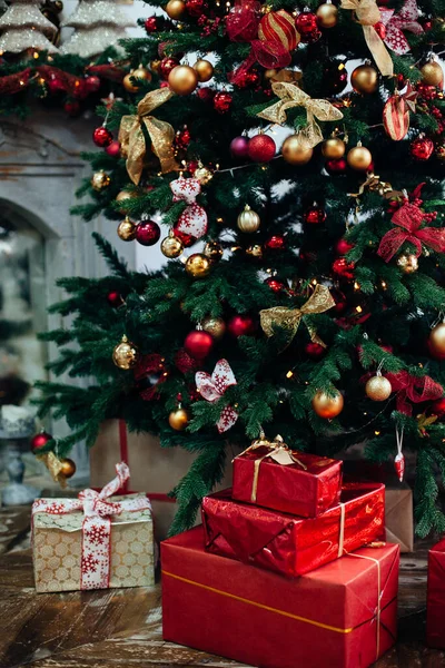 Stackar med julklappar i rött papper under en julgran dekorerade röda och gyllene bollar nära öppen spis i vardagsrummet.Nyår interiör — Stockfoto