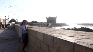  Genç Avrupalı mutlu kadın ve martı deniz kenarında Essaouira, Fas limanında arka planda uçan martılarla, ağır çekim