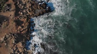 Üst manzara dalgaları mavi bir okyanusta kayalara çarpıyor. 4k 'lık deniz dalgalarının hava aracı görüntüsü. Kuş bakışı okyanus dalgaları yukarıdan boş bir taşa çarpıyor.