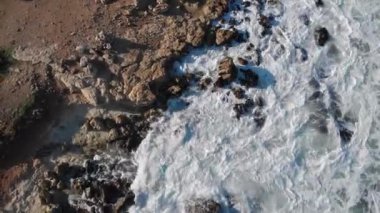 Yukarıdan bakıldığında gökyüzü dalgaları mavi bir okyanusta kayalara çarpıyor. İnsansız hava aracı deniz dalgaları 4K 'lık açıyla dönüyor. Dalgıç kıyafetli sörfçüler soğuk suda sörf yaparlar.