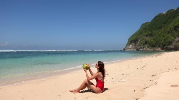 水の近くに座っている赤い水着の日焼けしたブルネットの女性、波を楽しんで、野生の熱帯ビーチ、バリ、インドネシアでココナッツジュースを注ぐ — ストック動画