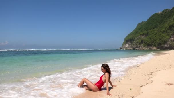 水の近くに座っている赤い水着の日焼けしたブルネットの女性、青い水で野生の熱帯ビーチで泡の波を楽しんで、バリ、インドネシア — ストック動画